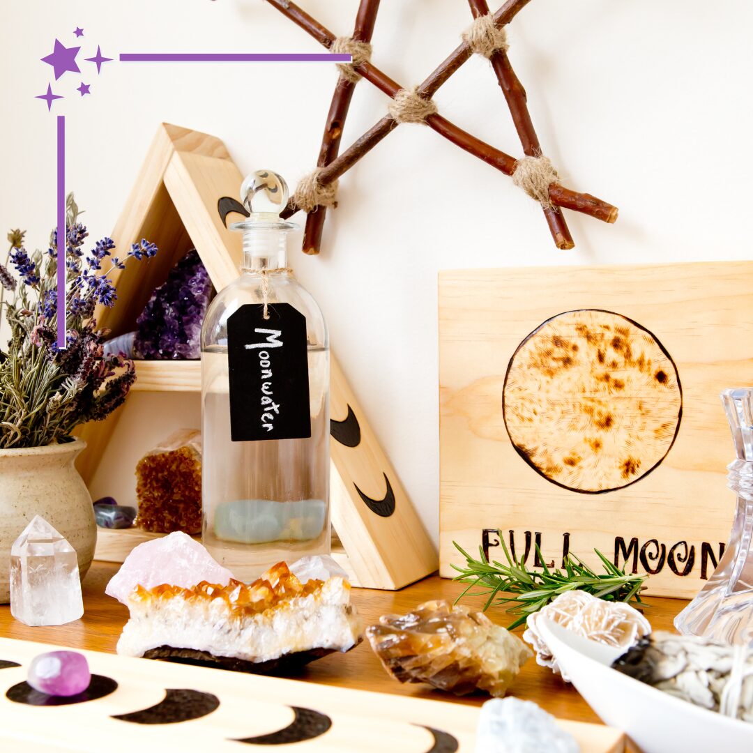 A tabletop display with a selection of crescent moon decorations, citrine, quartz and other crystals, a stoneware vase of lavender flowers, a jar of clear liquid labeled 'Moonwater,' a carved wooden display marked 'Full Moon', and a wood pentacle on the wall