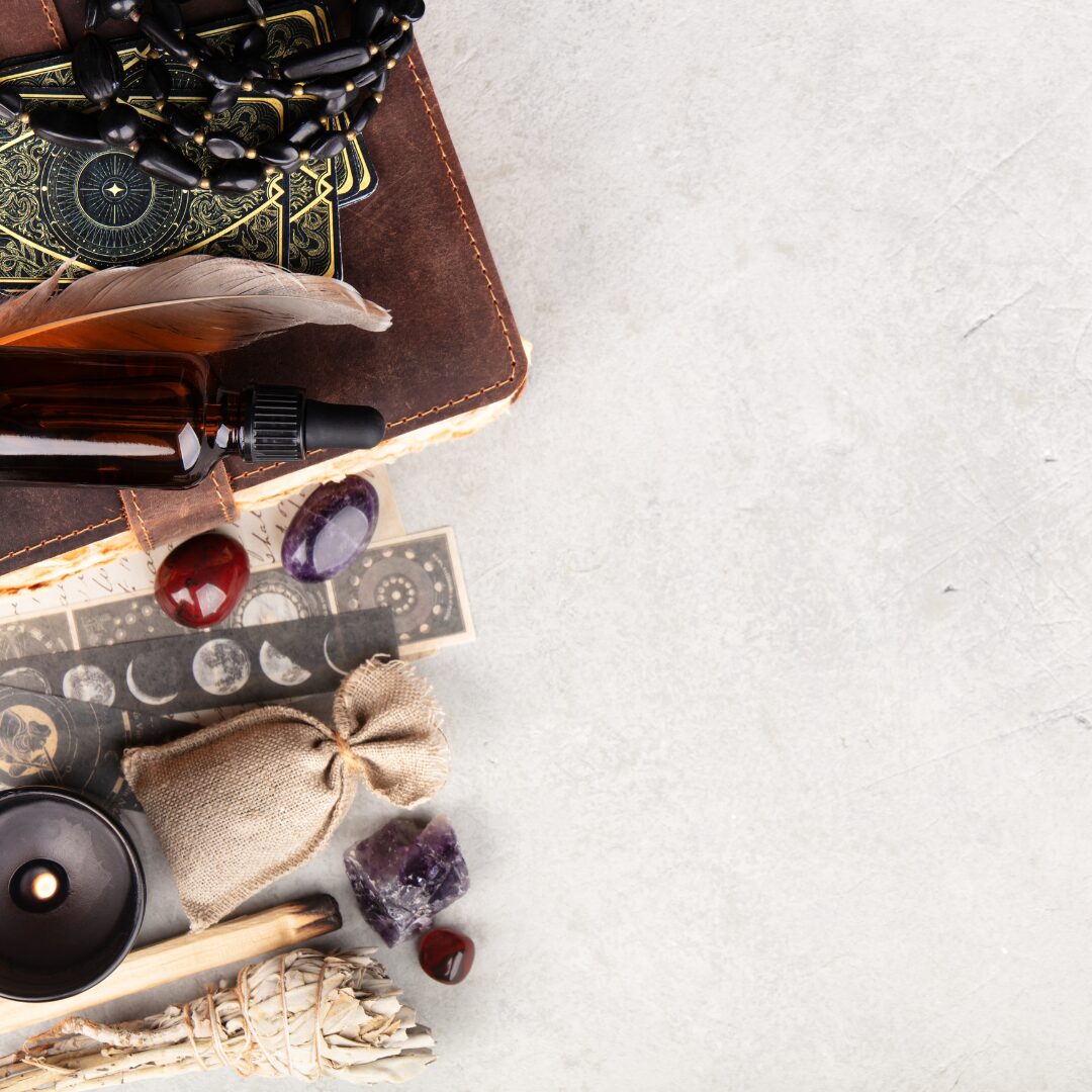 a selection of witchy supplies lined up, including a book, feathers, crystals, a candle, beads, sage bundle, burnt wood