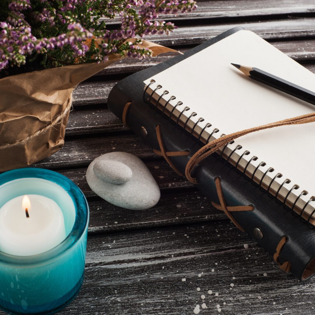 a burning candle, a bundle of flowers, and a pair of white stones sit next to a leatherbound book and a spiral notebook; the books stacked with a pen atop them.
