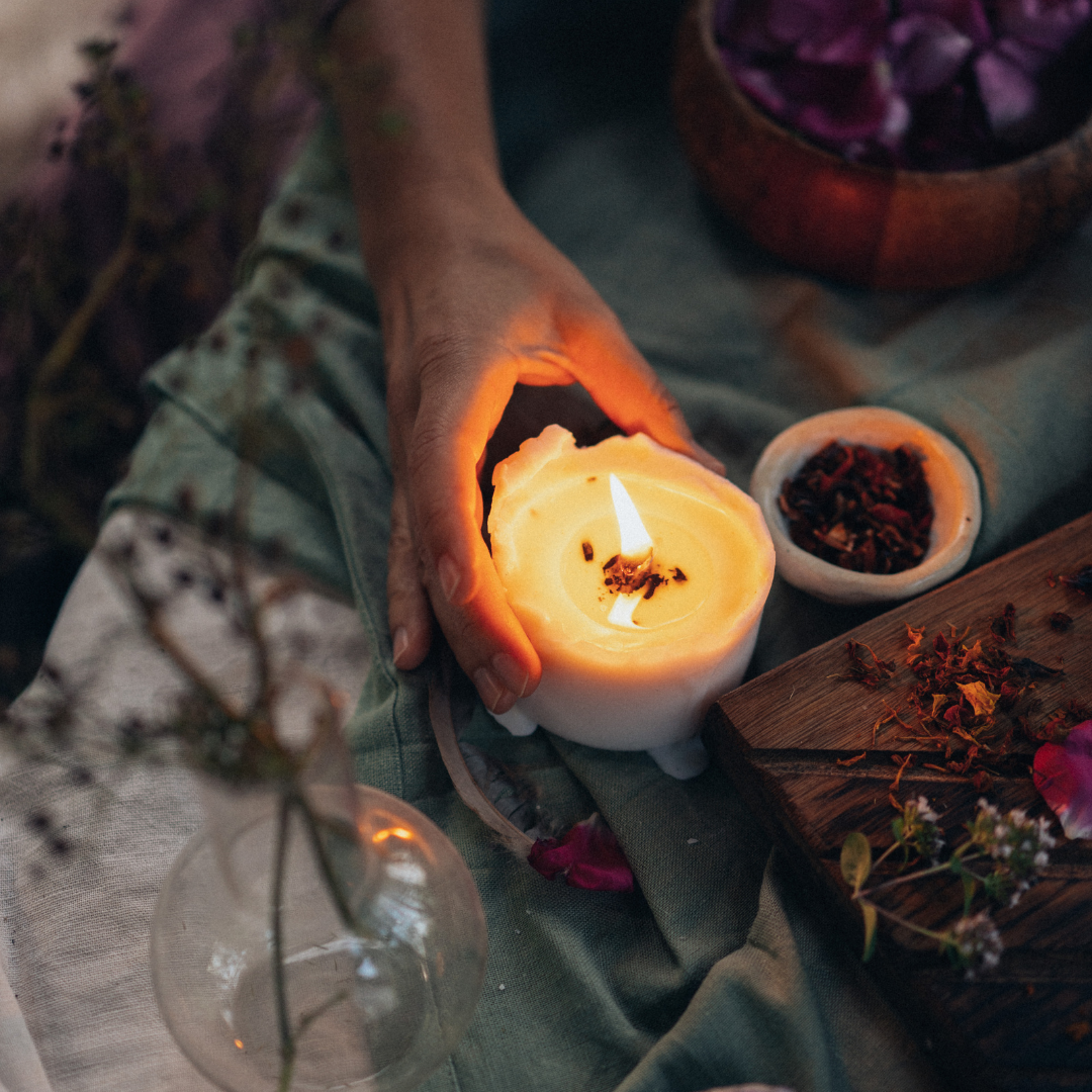 someone reaches for a lit candle, sitting near piles and small dishes of loose herbs and flowers