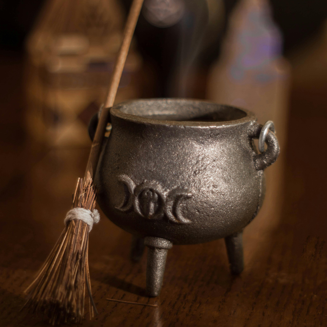 A miniature besom leans against a small cast iron cauldron embossed with the triple moon